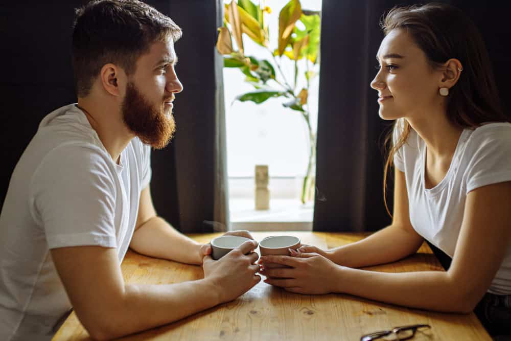 A man and a woman sit across from each other at a wooden table, holding mugs and talking, practicing lifestyle etiquette, with a plant and window in the background.