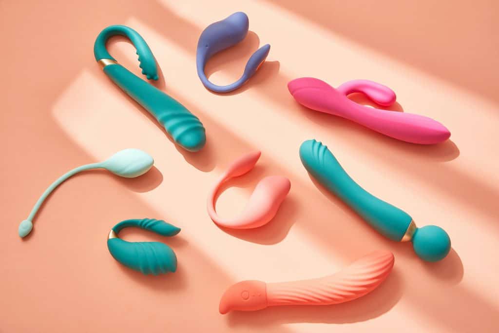 A variety of colorful silicone vibrators and best hotwife toys are arranged on a peach-colored surface with soft shadows.