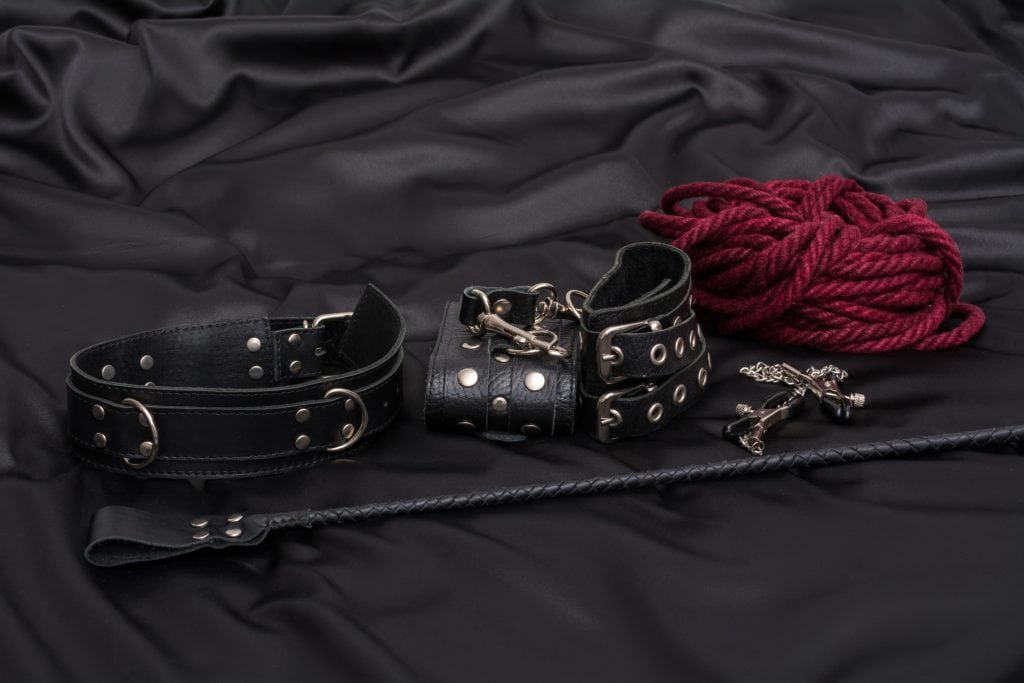Black leather collar, wrist cuffs, red rope, a small black whip, and a metal cross rest on black satin-a stylish nod to beginner BDSM and consensual kink exploration.